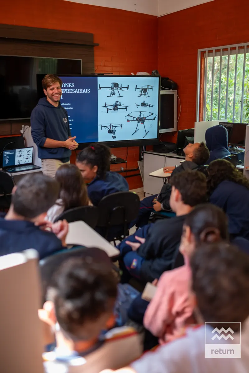 Em uma sala de paredes laranja, um instrutor está ao lado de uma tela com imagens de drones. Jovens sentados em cadeiras acompanham a apresentação.