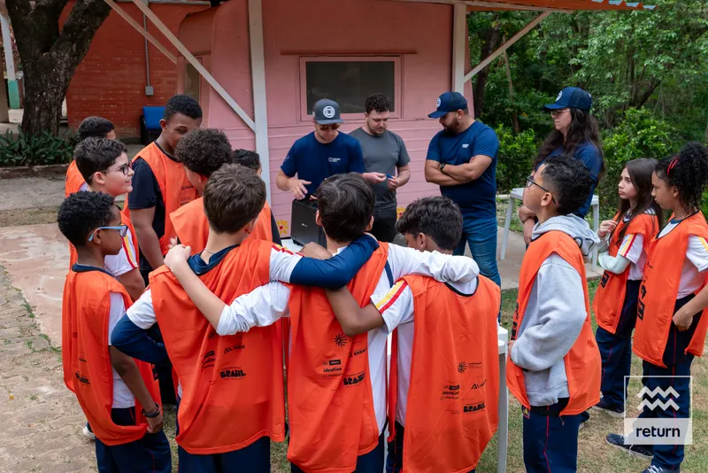 Jovens com coletes laranja formam uma roda ao ar livre. Alguns estão abraçados pelos ombros. Instrutores de camisetas azuis conversam com o grupo, junto a uma construção rosada.