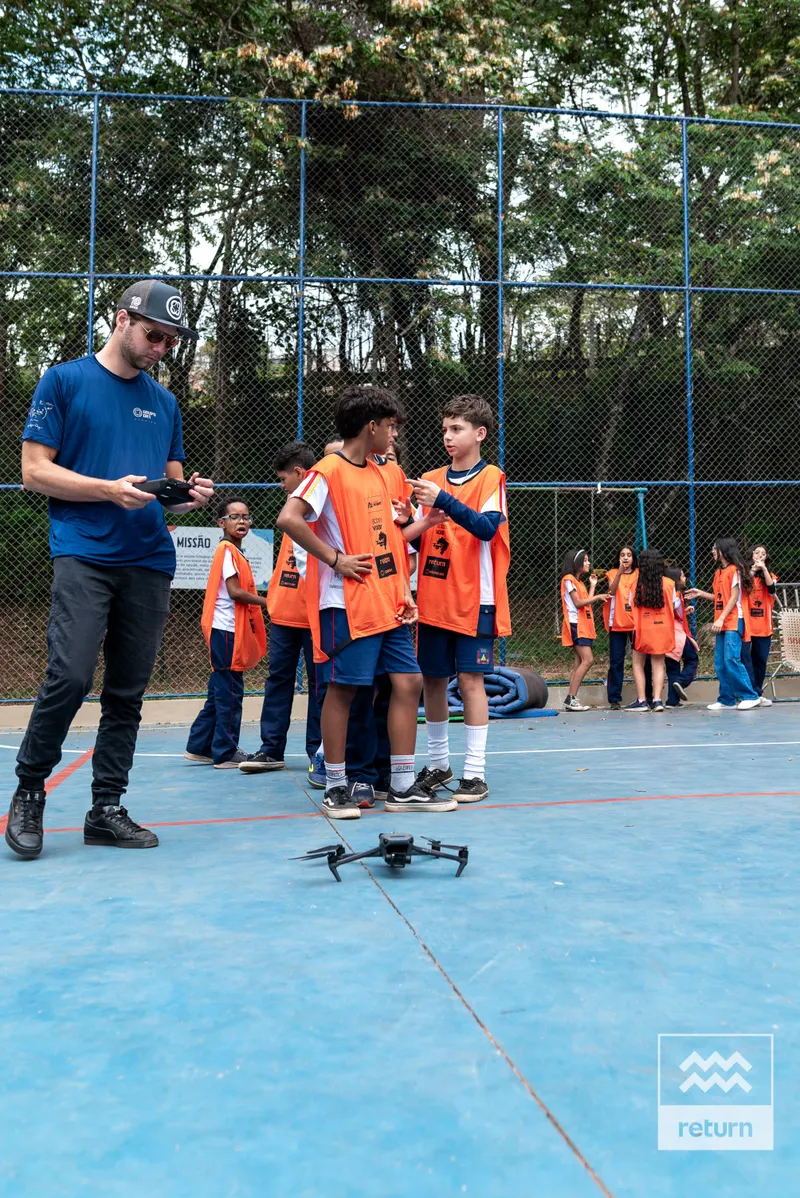 Em uma quadra azul cercada por alambrado e árvores, um instrutor acompanha jovens com coletes laranja. Um drone repousa no chão à frente do grupo.