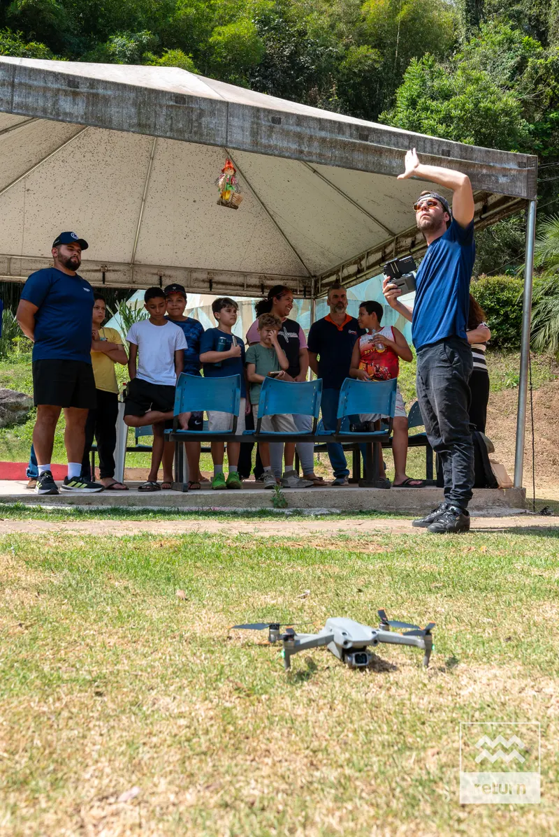 Um drone está sobre o gramado em primeiro plano. Ao fundo, jovens se reúnem sob uma cobertura, enquanto um instrutor ao lado ergue um dos braços.