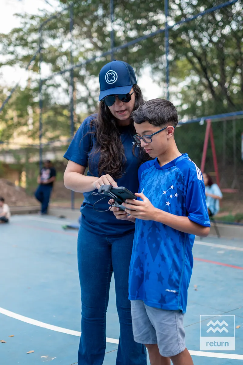 Na quadra, uma instrutora de boné e camiseta azul acompanha um jovem de óculos e camiseta azul que segura o controle de um drone.