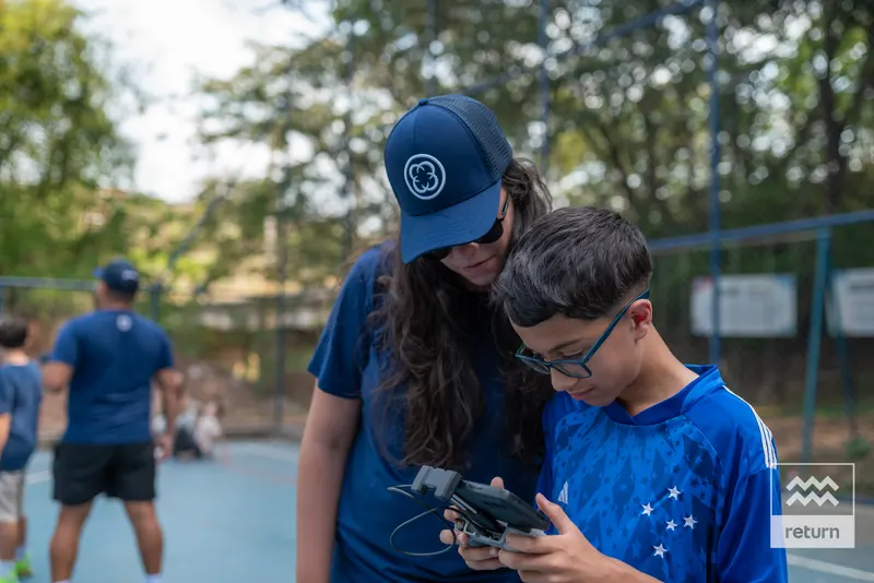 Uma instrutora de boné azul se inclina ao lado de um jovem de óculos. Os dois olham para o controle que ele segura, com a quadra e outros participantes ao fundo.