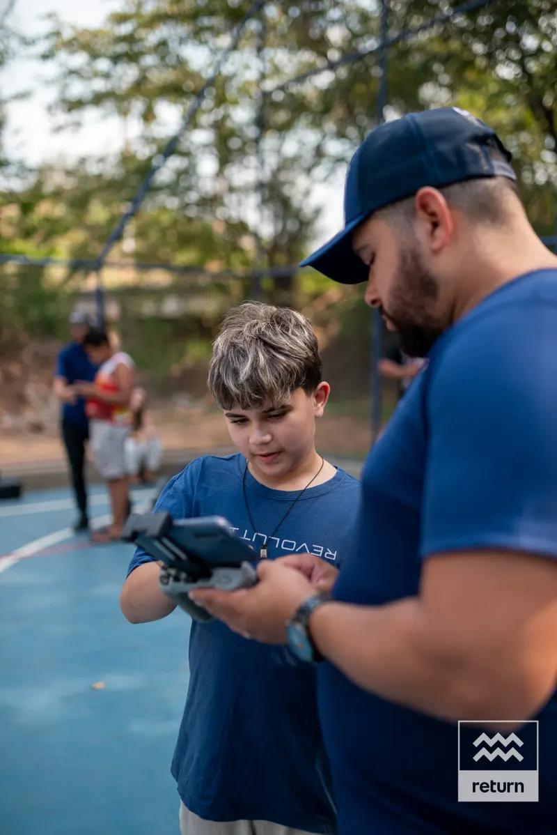 Um jovem de camiseta azul olha para o controle que segura com as duas mãos. Ao seu lado, um instrutor de boné acompanha a atividade na quadra.