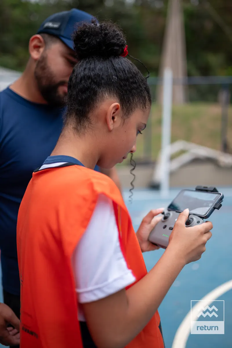 Visto de lado, um jovem com colete laranja segura um controle com tela. Um instrutor de camiseta azul está logo atrás dele, acompanhando a operação.