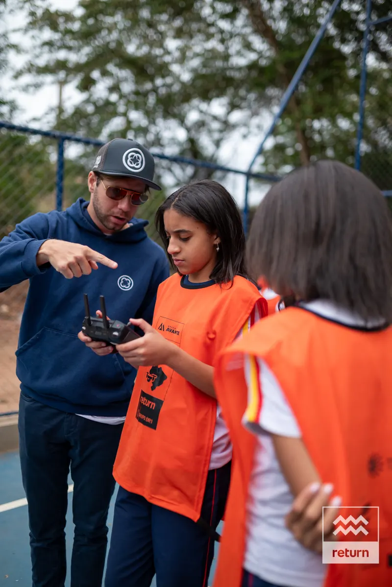 Um instrutor de boné aponta para o controle segurado por uma jovem de colete laranja. Outra participante acompanha a explicação na quadra cercada por árvores.
