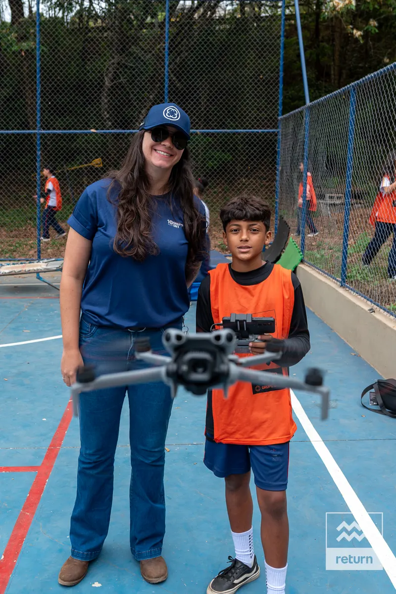Uma instrutora e um jovem de colete laranja estão lado a lado na quadra. Um drone aparece em destaque à frente deles; o jovem segura um controle.