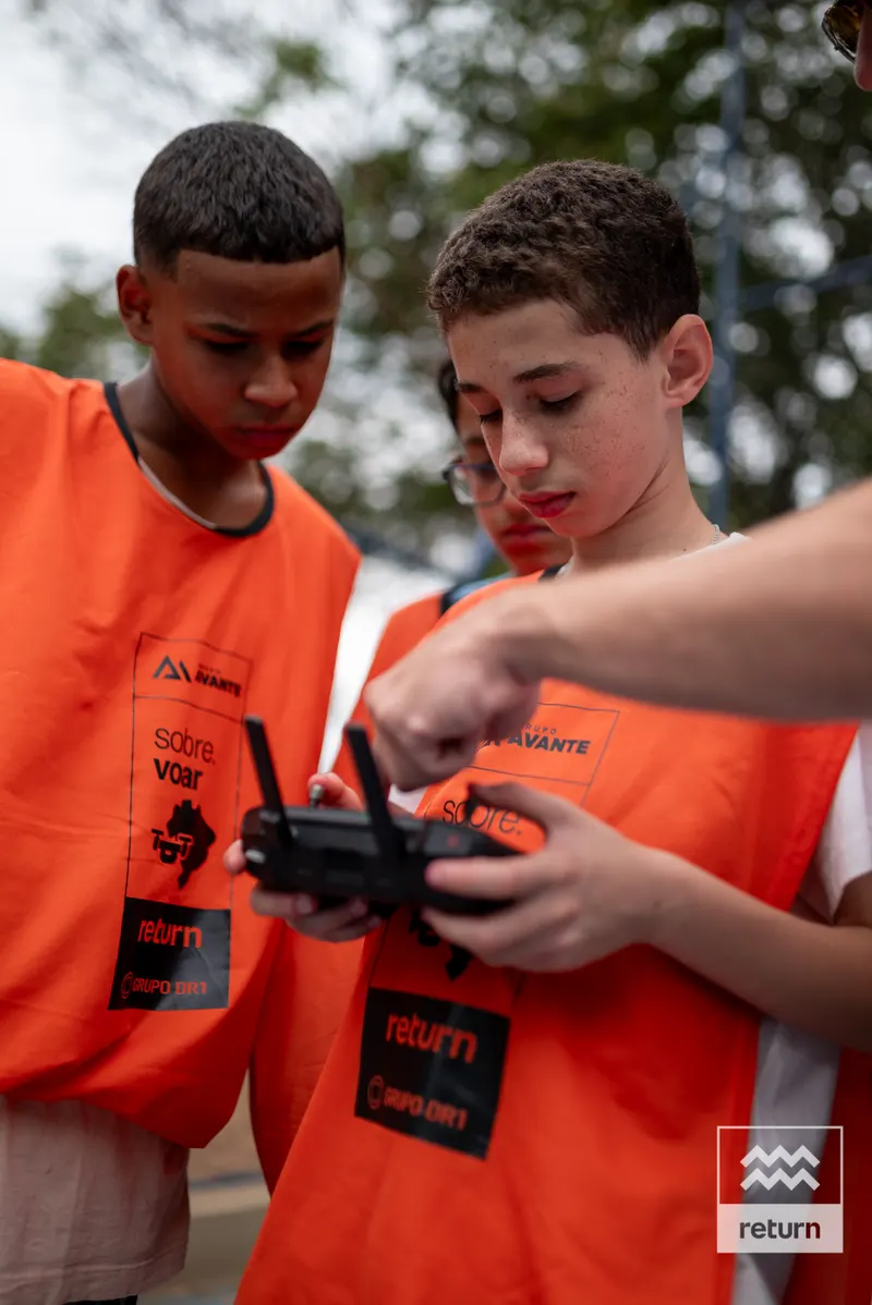 Dois jovens de coletes laranja observam um controle. As mãos de um instrutor entram pela direita e apontam para os comandos do equipamento.