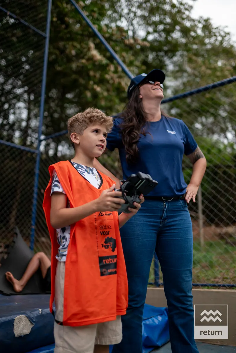 Um jovem de colete laranja segura o controle de um drone e olha para cima. Uma instrutora ao seu lado também observa o alto, com árvores ao fundo.