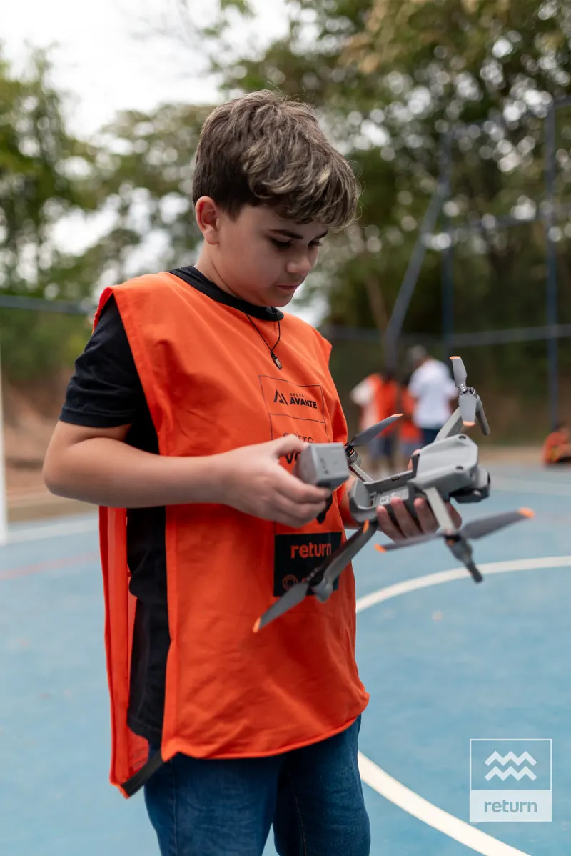 Na quadra azul, um jovem de colete laranja olha para um drone e um controle que segura próximo ao corpo. O alambrado e as árvores aparecem ao fundo.