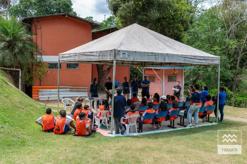 Participantes estão reunidos sob uma tenda branca ao lado de uma construção rosada. Alguns se sentam em cadeiras e outros no gramado, em meio a árvores.