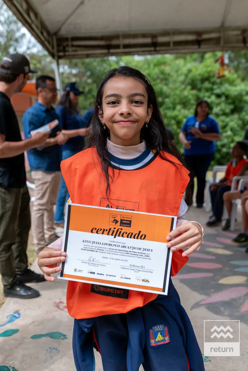Uma jovem de colete laranja está de frente para a câmera, segurando um certificado com moldura laranja. Outros participantes aparecem ao fundo, sob uma cobertura.