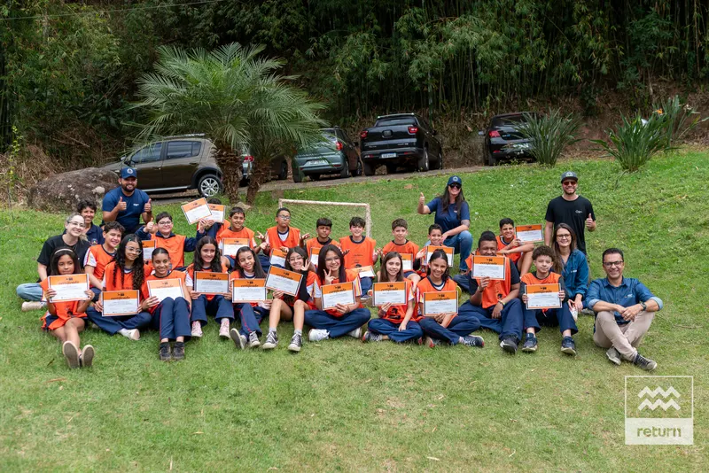 Jovens de coletes laranja e adultos posam em um gramado, organizados em fileiras, segurando certificados. Árvores e veículos aparecem atrás do grupo. A marca Return está no canto inferior direito.